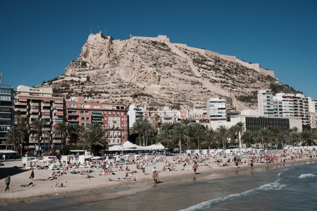 A view of Alicante’s Playa del Postiguet with clear blue waters, golden sand, and the historic Santa Bárbara Castle in the background.