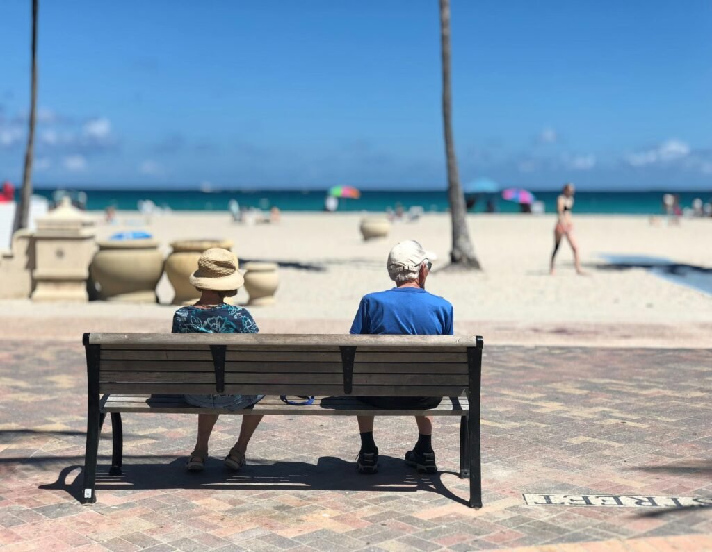 US military retirees living overseas enjoying retirement: An older couple sitting on a bench by the beach in a warm, peaceful destination.